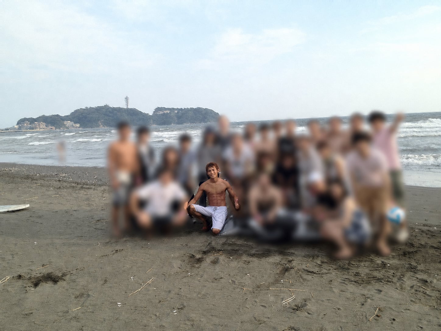 曇り空の砂浜での集合写真。奥に江ノ島と展望灯台のシルエット、打ち寄せる波。波打ち際に大勢が並び、手前中央にひとり片膝をついて座っている