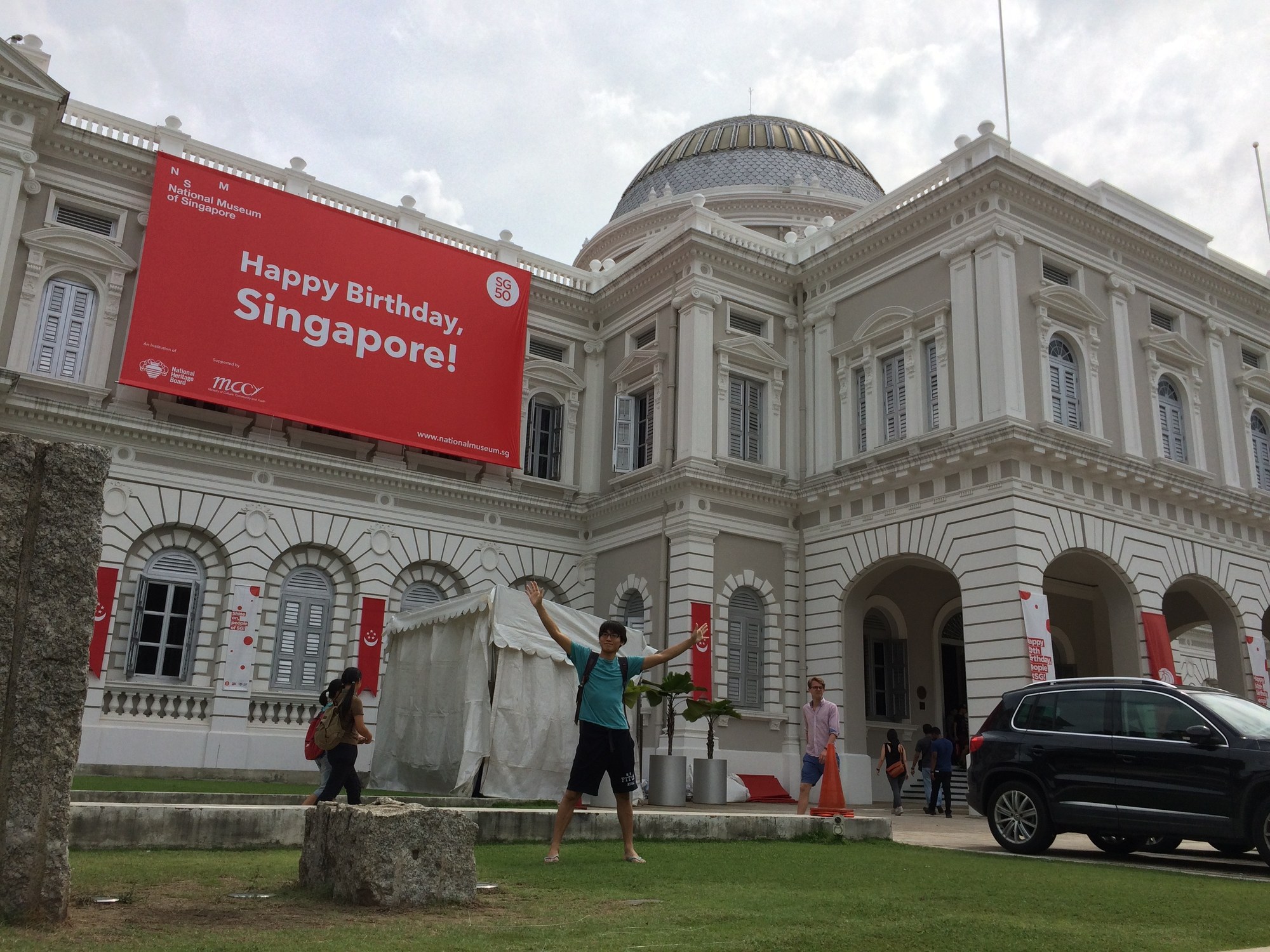 シンガポール国立博物館の白い建物。正面に「Happy Birthday, Singapore!」の赤い横断幕が掛かり、芝生の手前で腕を上げて立っている