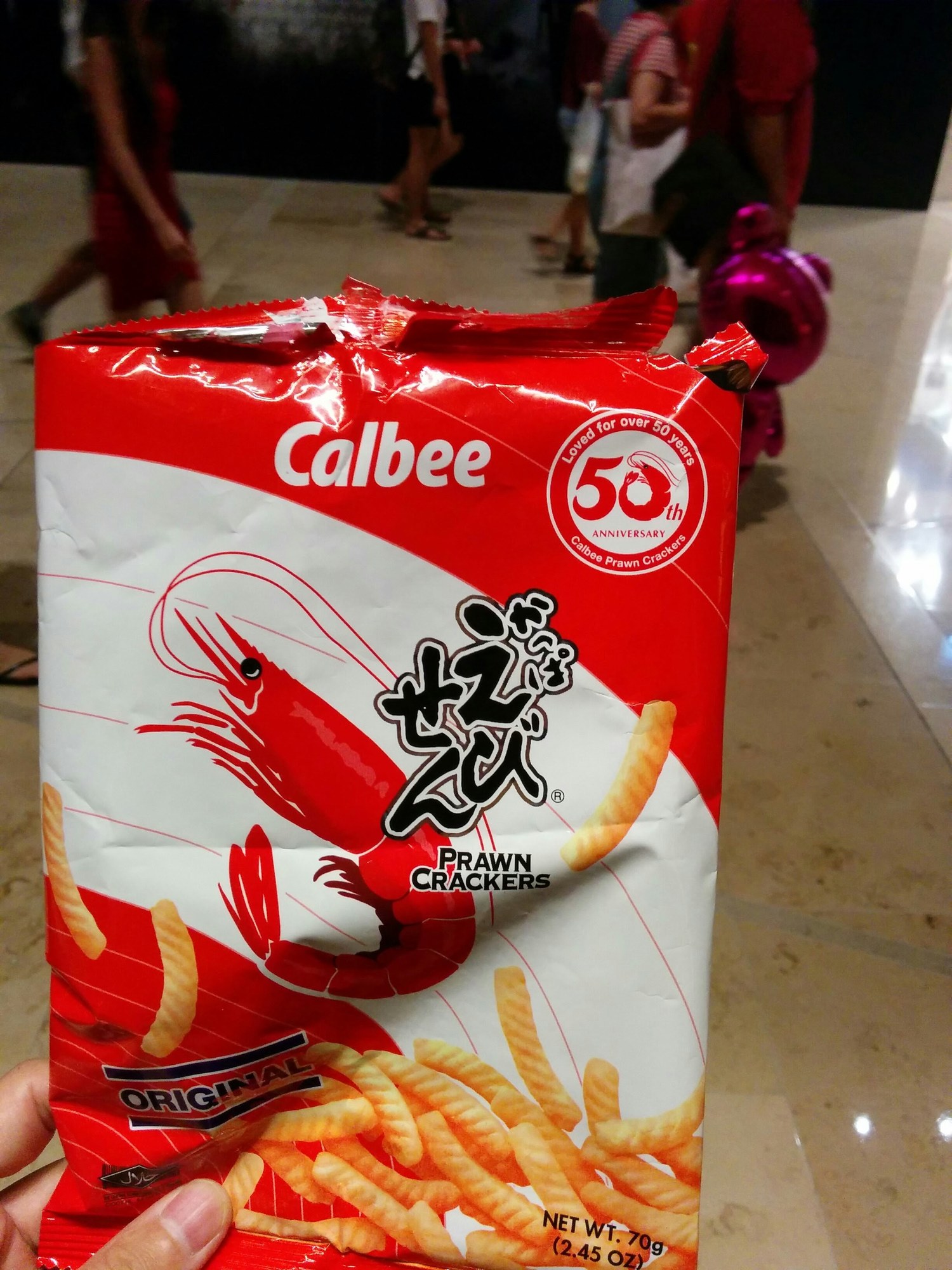 Calbee のかっぱえびせん（PRAWN CRACKERS・ORIGINAL・70g）の袋。右上に「Loved for over 50 years / 50th ANNIVERSARY」のロゴ。奥のマリーナベイの通路を赤い服の人たちが行き交っている