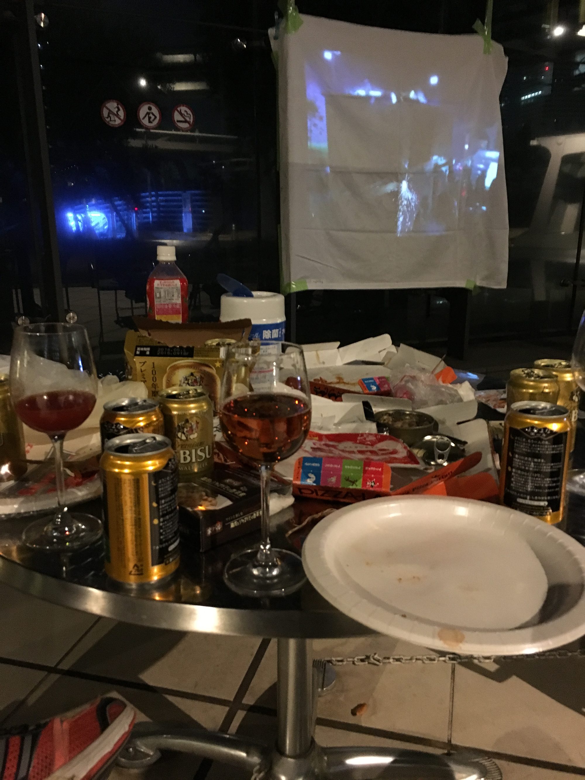 白い布を貼った即席スクリーンに映画を投影。テーブルにはビール缶やワイングラス、ピザの箱やスナックが散らかり、窓の外は夜景