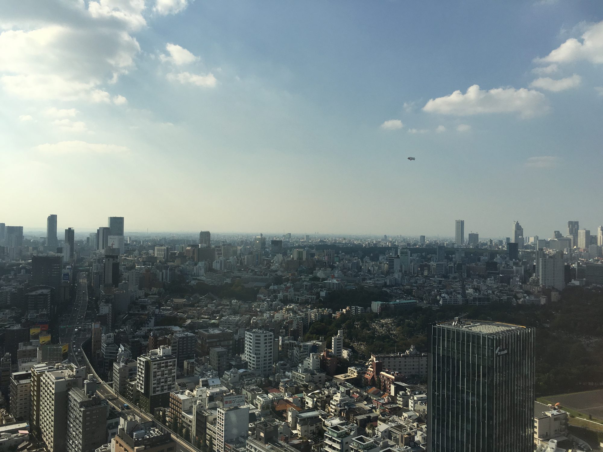 高層オフィスから見た東京の街並み。かすんだ空に小さな飛行船が浮かぶ