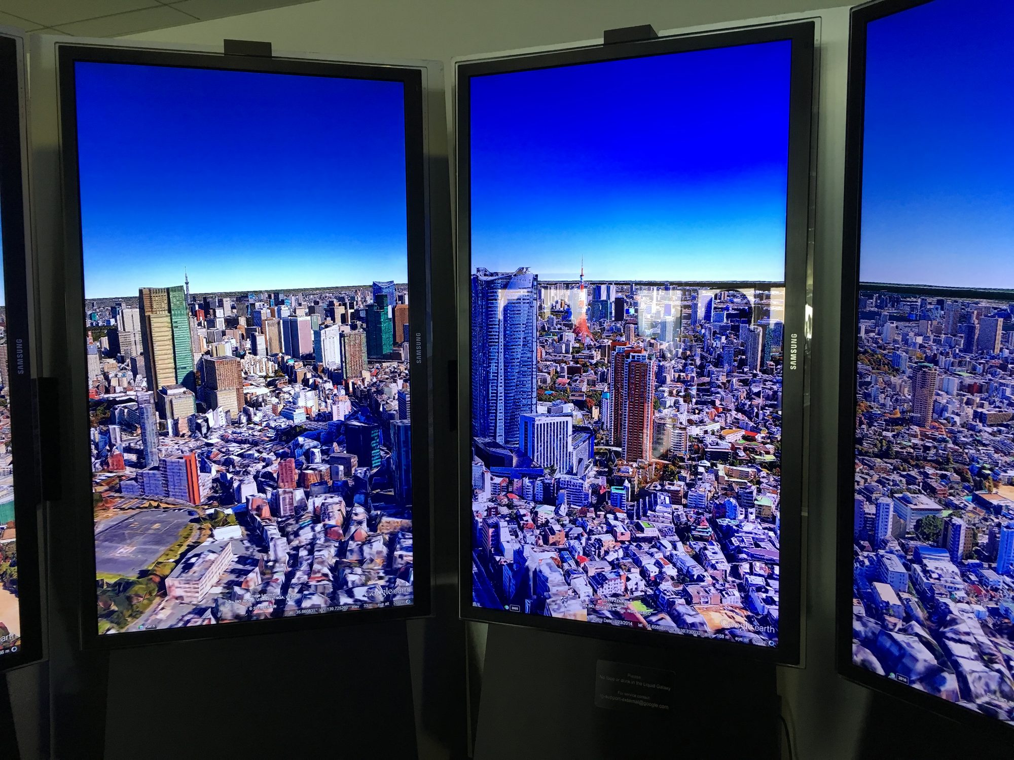 Google 六本木オフィスの Liquid Galaxy。縦置きの Samsung ディスプレイ数台に Google Earth の東京3D（東京タワーが見える）が映る