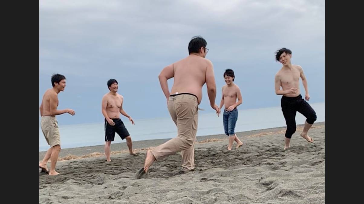曇り空の砂浜で、上半身裸のエンジニアたちが相撲をとって遊んでいる。中央の一人が突進する