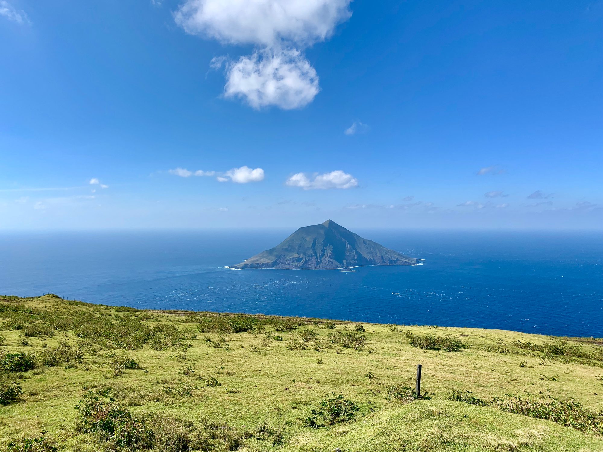 草原の丘の先、紺碧の海に浮かぶ円錐形の八丈小島。快晴の空
