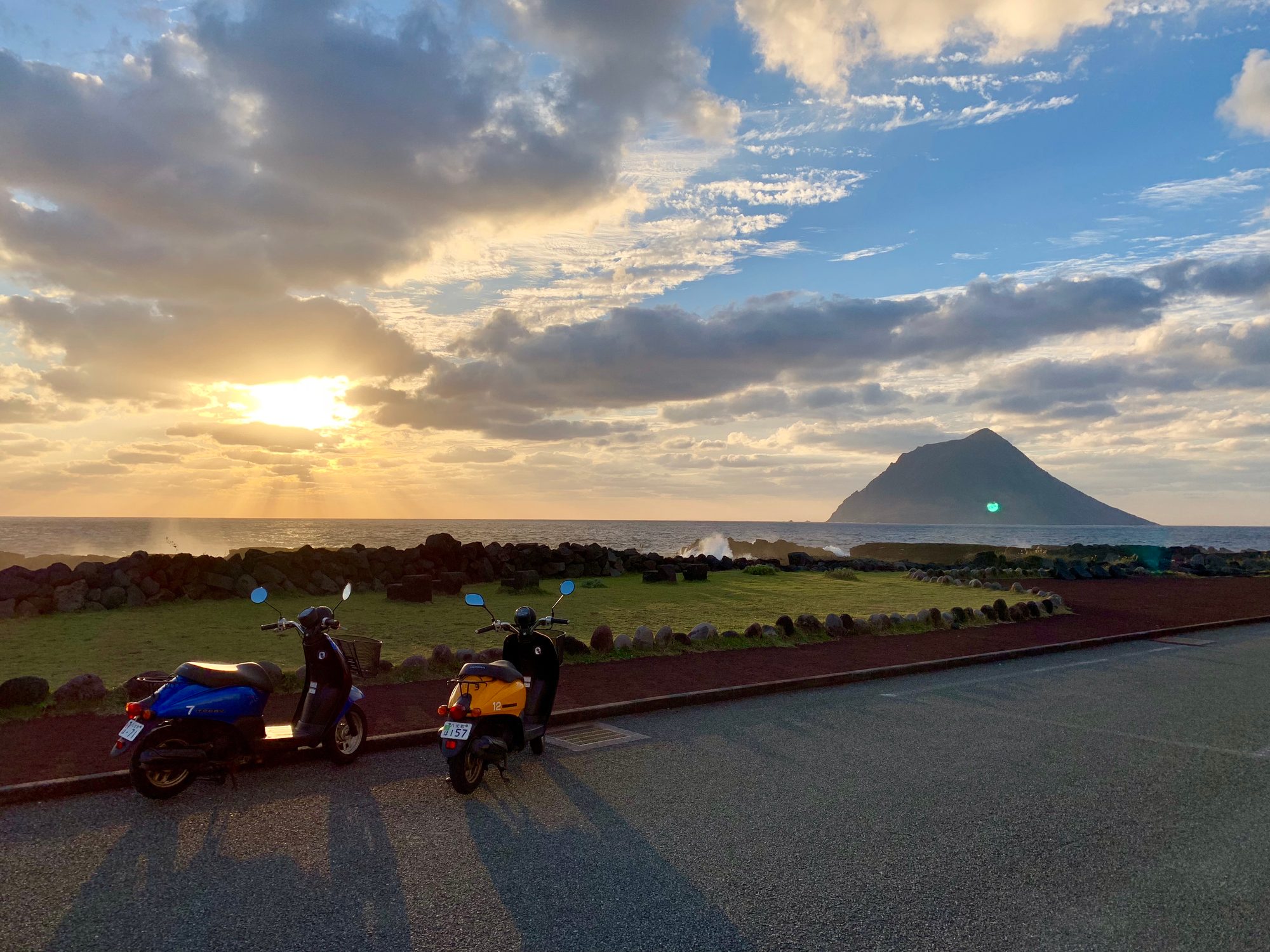 夕暮れ、石垣のある草地の道路に停めた青と黄のスクーター2台。海の向こうに八丈小島のシルエットと沈む夕日