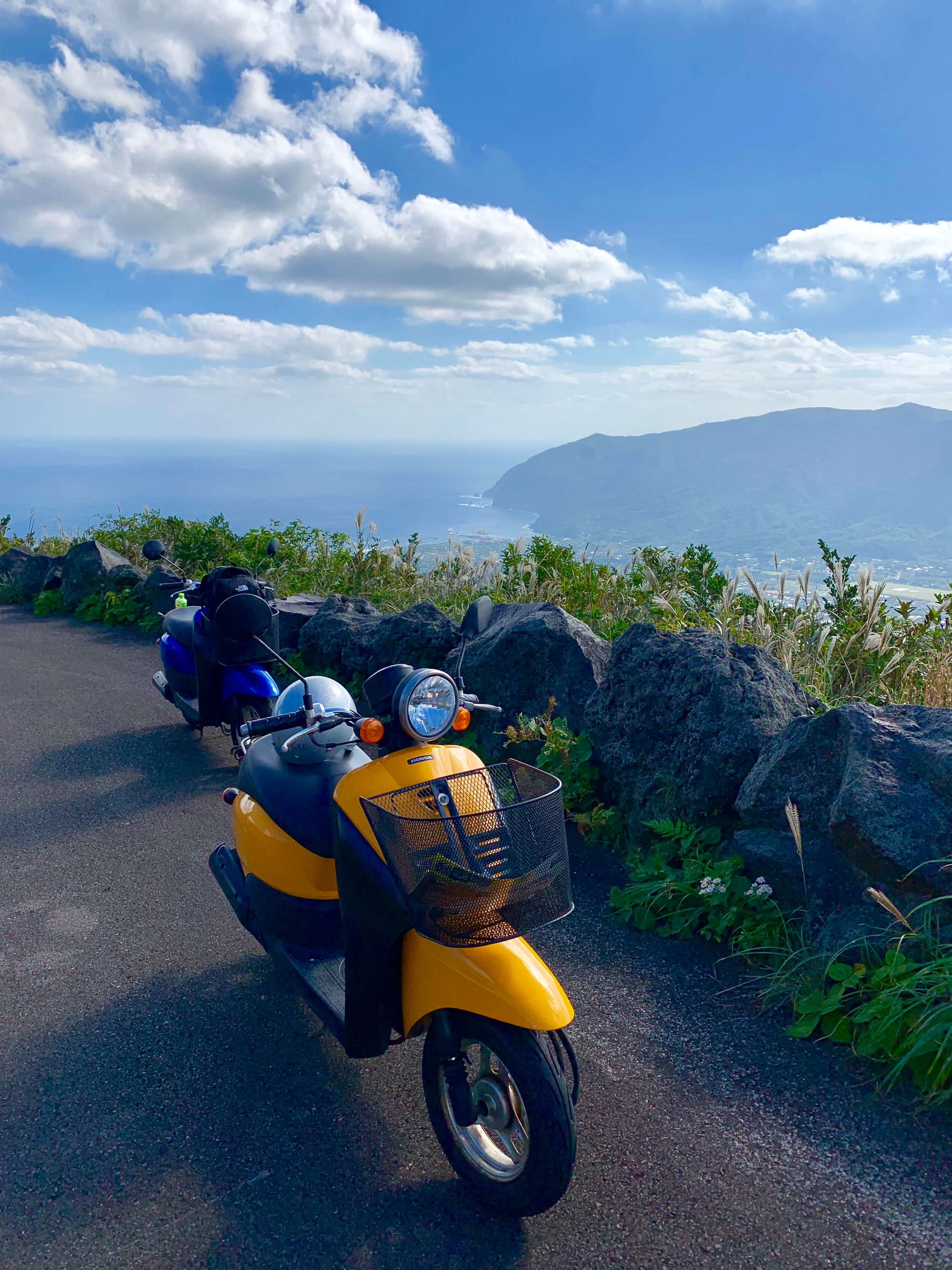 展望台の道路に停めた黄色と青のレンタルスクーター2台。眼下に八丈島の海岸線と山、青空が広がる