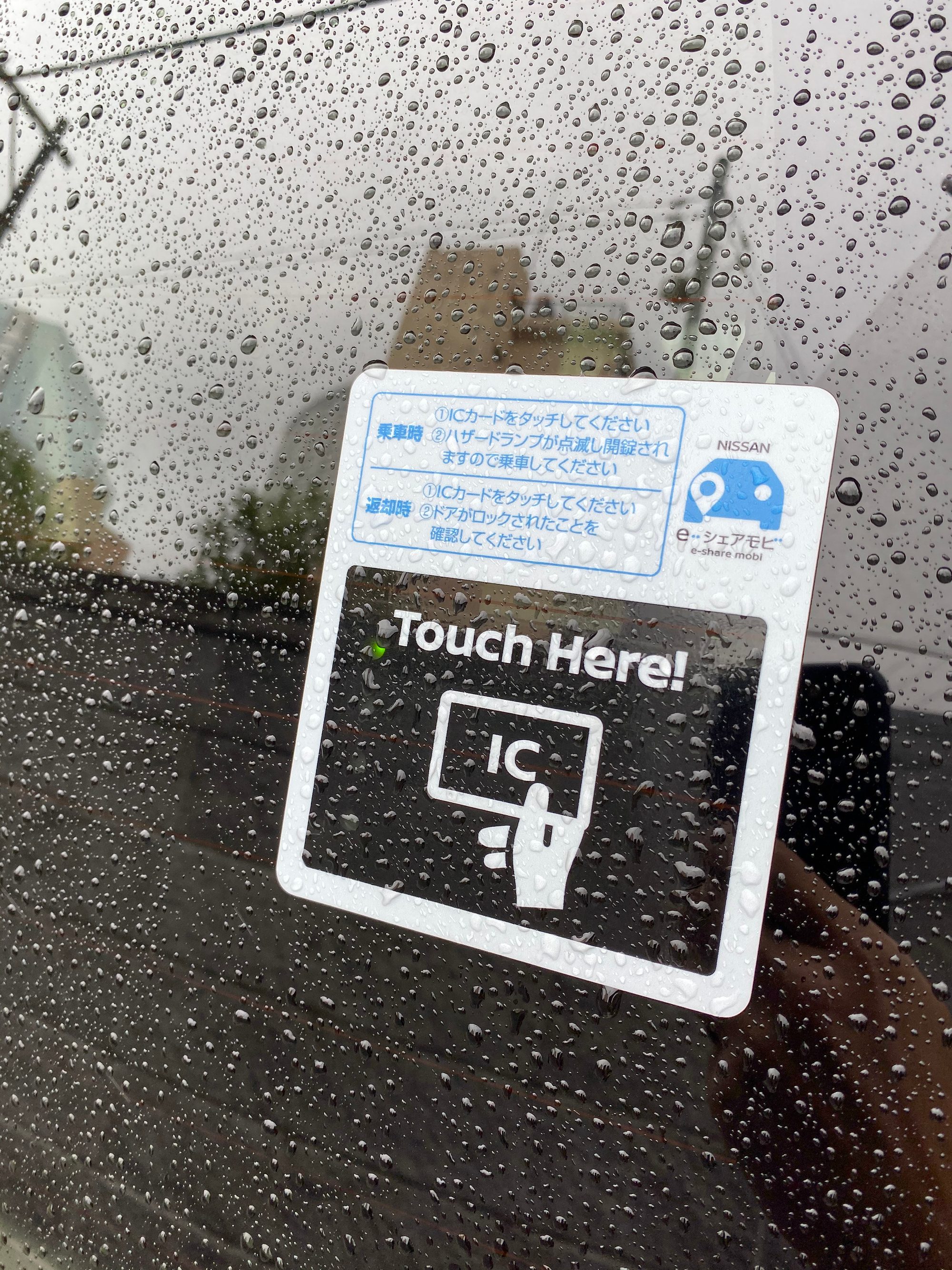 カーシェア（日産 e-シェアモビ）の車の窓に貼られた「Touch Here!」IC カードの案内ステッカー。雨粒が付いている