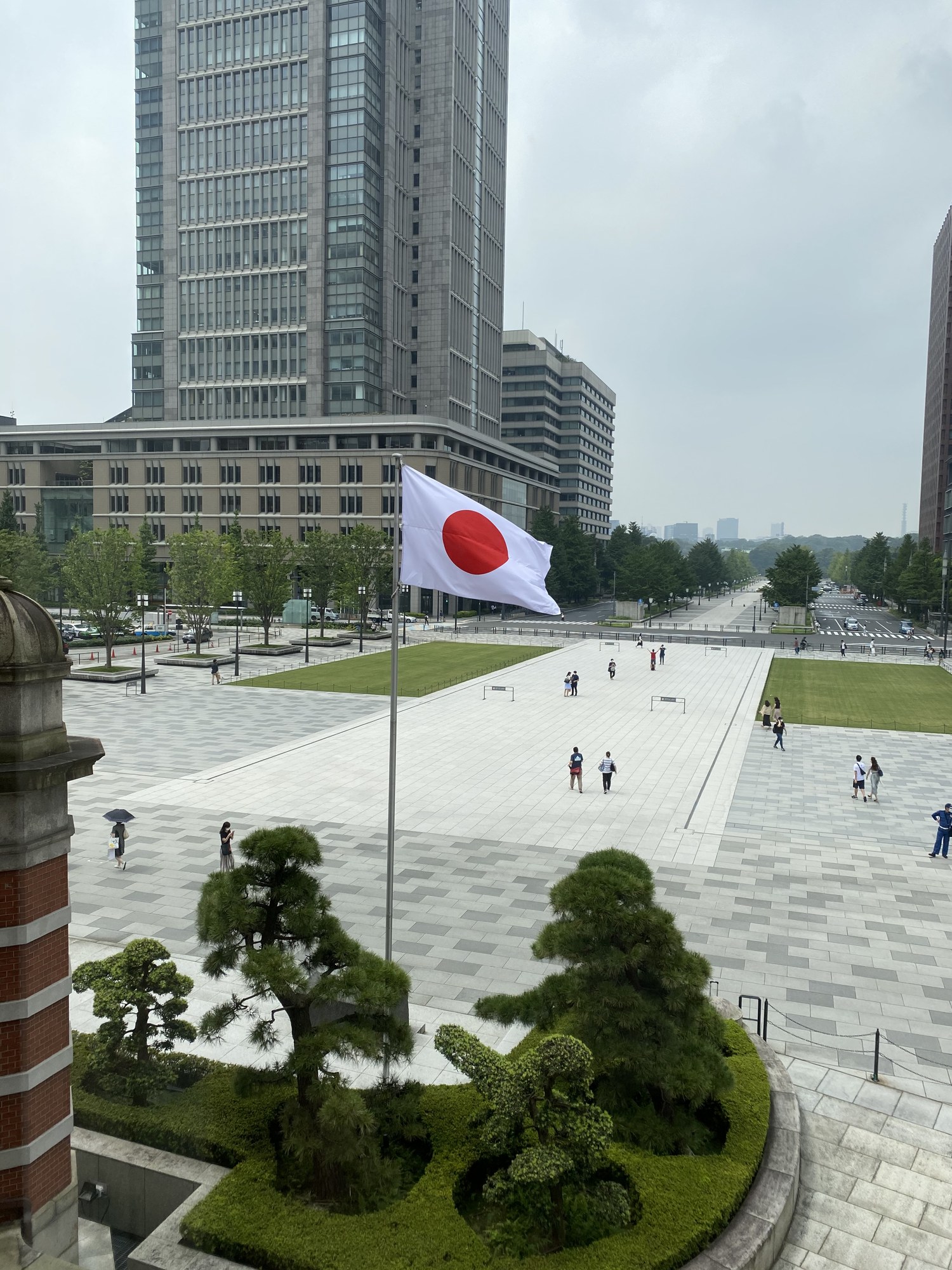 東京駅丸の内側の駅前広場。日章旗がはためき、その先に行幸通りが伸びる。手前に松の植え込み、奥に丸の内のビル群