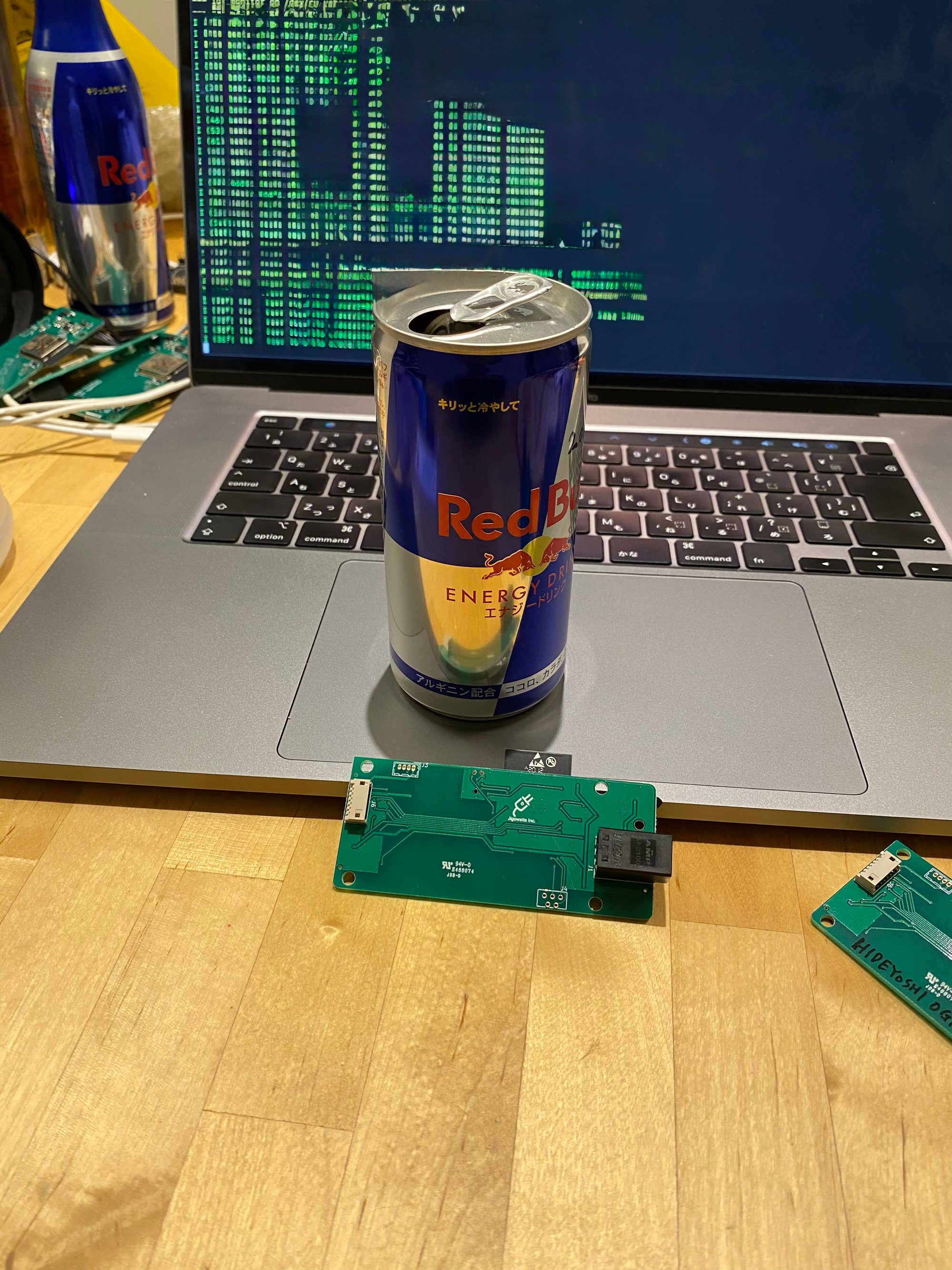 自動車向けスマートロックの開発風景。MacBook の画面にマトリックス風の緑の文字、トラックパッドに Red Bull、机に自作の緑の基板（「HIDEYOSHI OGA」と書かれたものも）