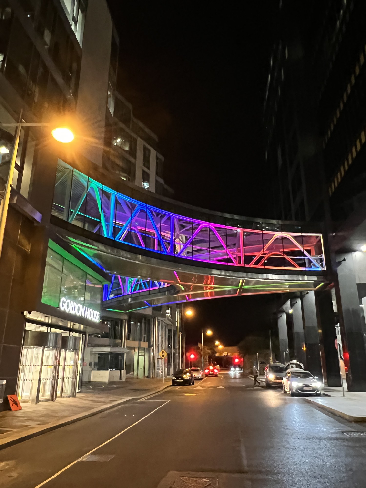 夜の通りをまたぐ、虹色にライトアップされた連絡橋。手前に Gordon House の建物と街灯