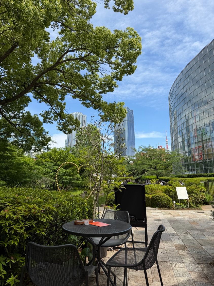 毛利庭園のオープンカフェ。黒いテーブルと椅子の向こうに新緑の木々、テレビ朝日のガラス張りの建物と高層ビル、青空